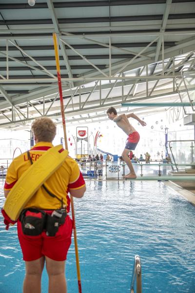 Boy jumping off small dive board with lifeguard in foreground