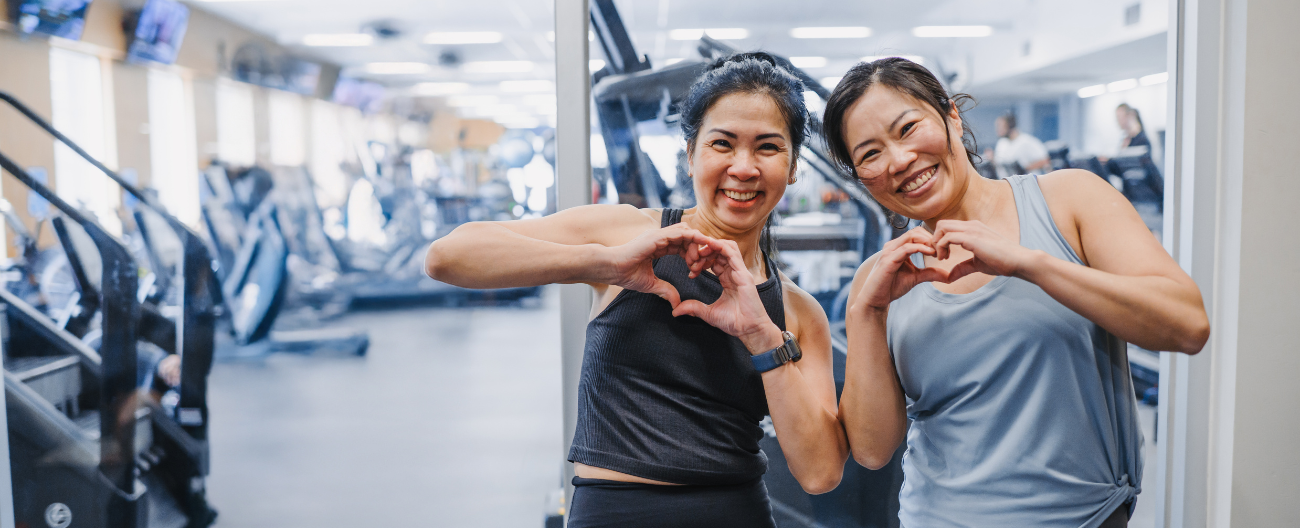 Image of two women smiling in gym, both making love heart shapes with their hands.