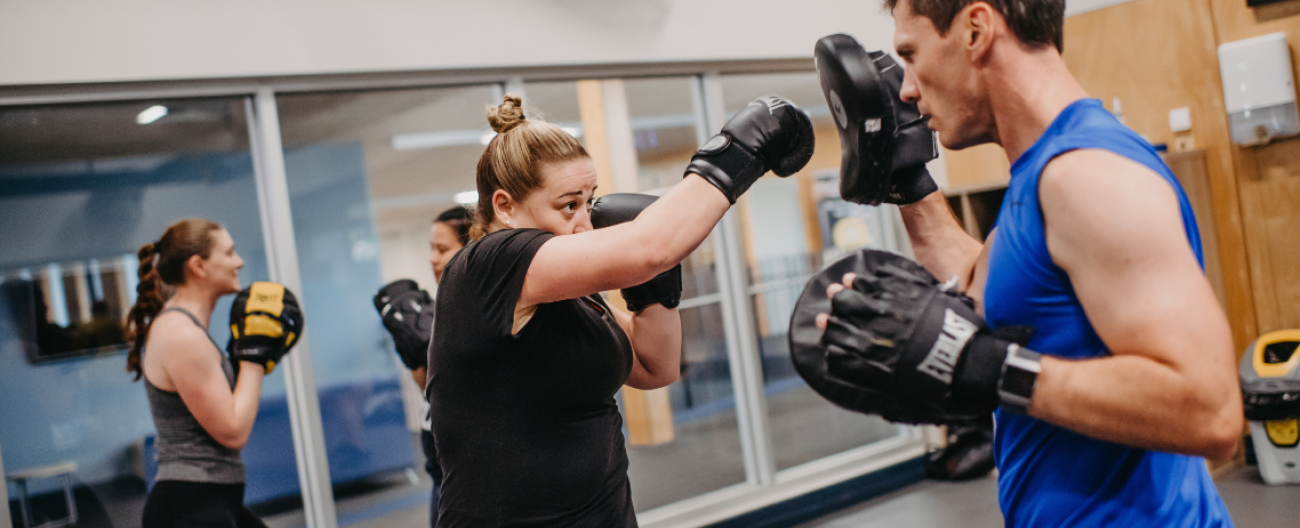 two people in a boxing class, man in blue singlet holding pads as person in black top throws a punch